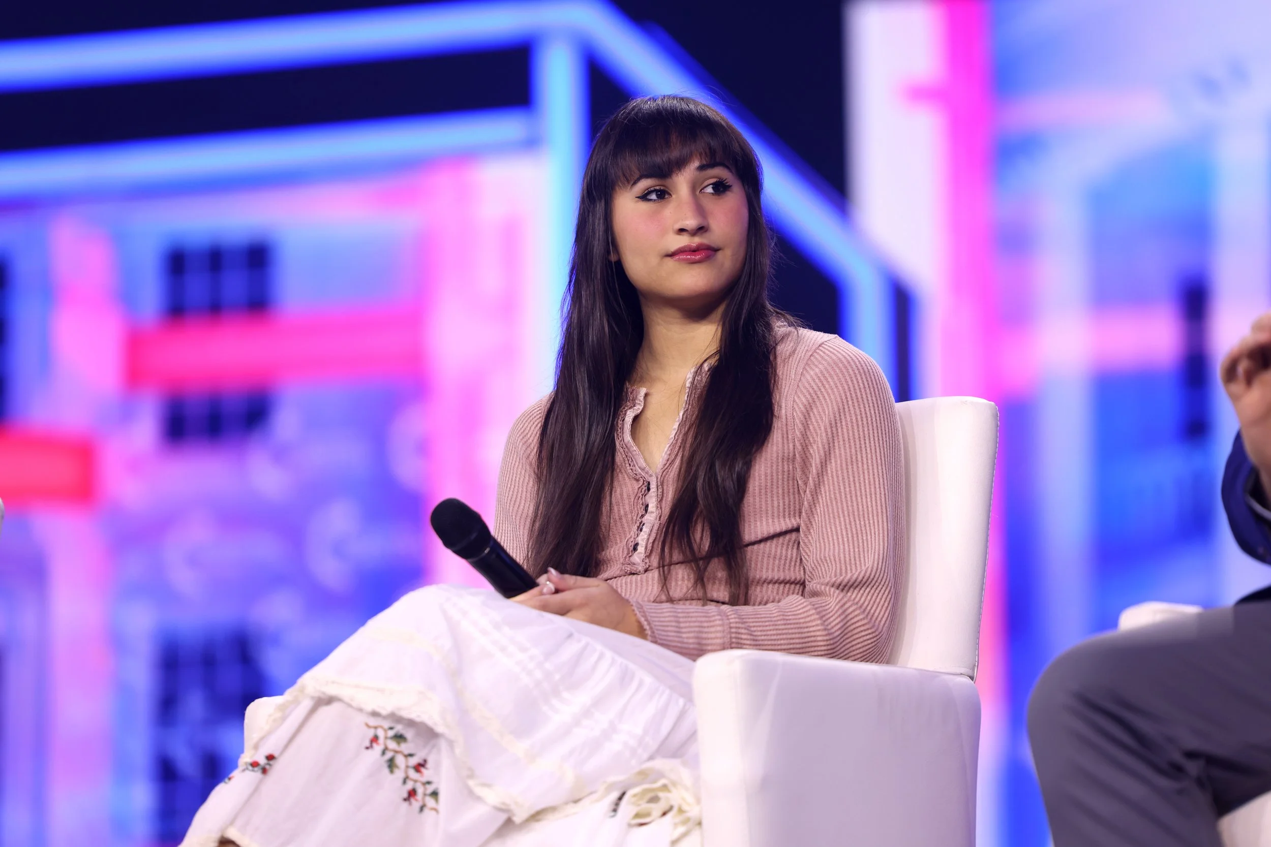 Chloe Cole seated on a pink and blue lit stage with a microphone